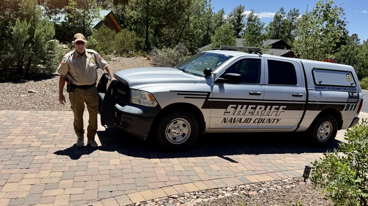 Uniformed Navajo County Sheriff's Auxiliary volunteer standing beside a marked Sheriff Auxiliary patrol truck in a pine-lined neighborhood driveway